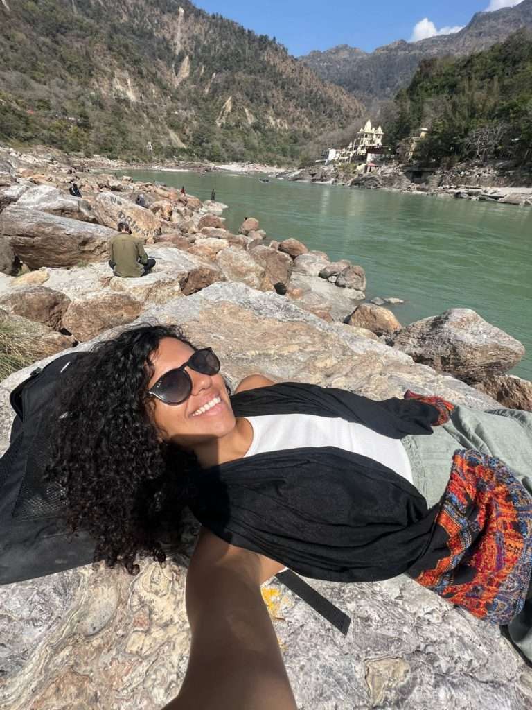 A woman with curly hair wearing sunglasses and a white top lies smiling on rocks with water and mountains in the background. She has a colorful fabric accessory around her waist and reaches out to take the photo in Rishikesh India