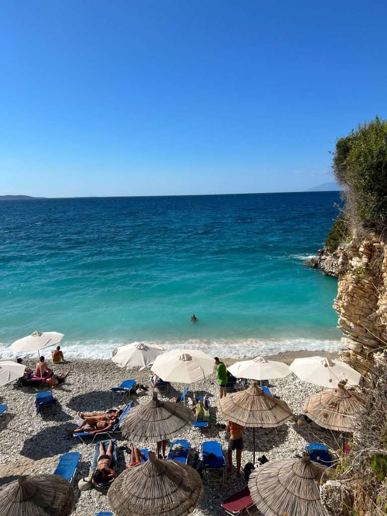 A picturesque beach with turquoise waters and white pebbles awaits on your 1-week Albania itinerary. People relax under straw umbrellas on lounge chairs, enjoying the sunny weather. One person swims in the sea, while the clear blue sky stretches overhead. A rocky formation is visible on the right - Pulëbardha beach