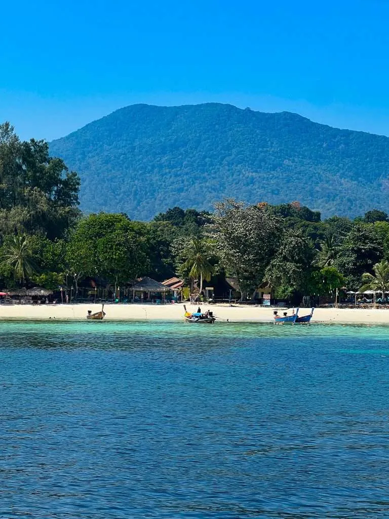 A serene tropical scene of Pattaya Beach In Koh Lipe, Thailand lined with trees and small huts offers one of the best things to do in Koh Lipe. Several boats float on the clear blue water, and a majestic, wooded mountain rises under a clear sky, providing a stunning backdrop for unforgettable adventures.