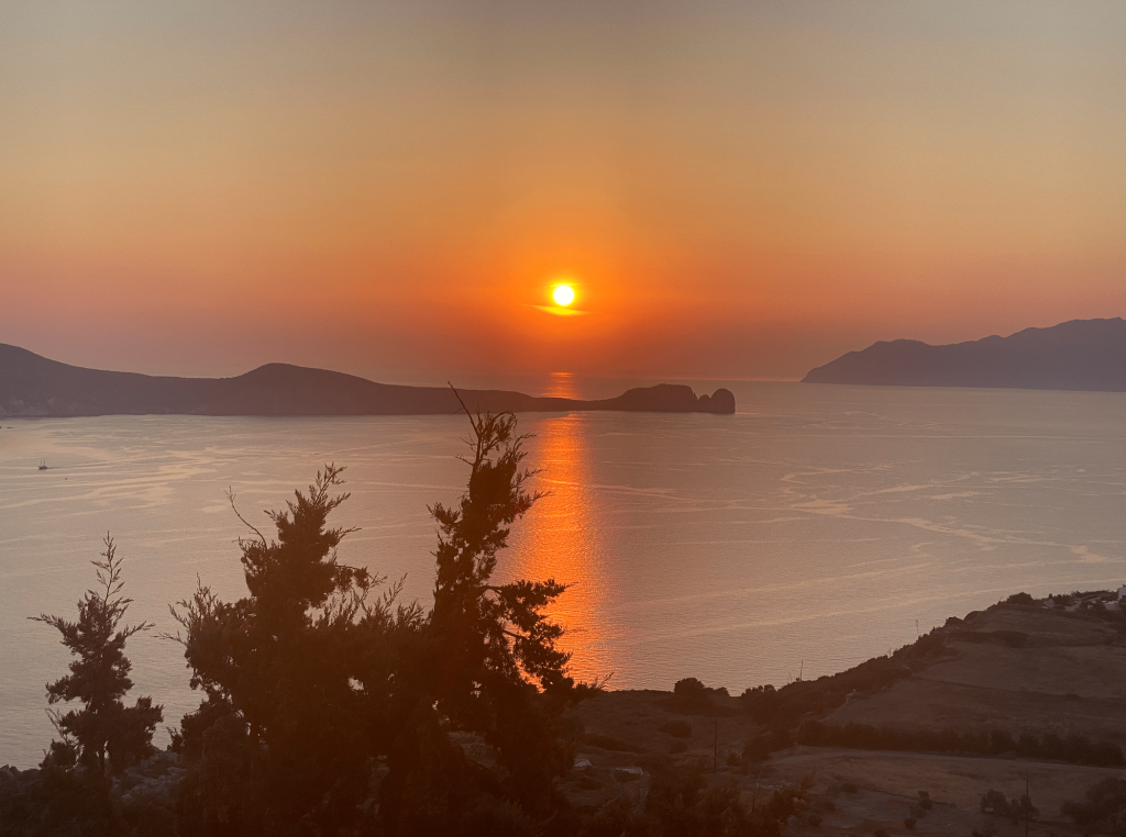 A picturesque sunset over a calm sea with the sun low in the sky and its reflection creating a golden path across the water. Silhouetted trees and a distant island are visible, with gentle hills framing the tranquil scene, just one of many breathtaking views on our 5-Day Greece Itinerary - View from Utopia Bar in Plaka Milos