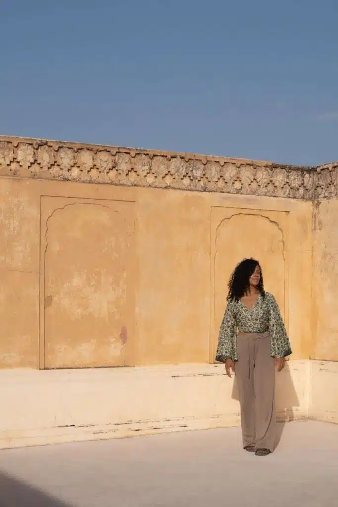 A woman with curly hair, wearing a patterned green top and beige pants, stands on a sunlit rooftop with textured yellow walls—evoking the question: Is India Safe For Solo Female Travellers—gazing to her left under a clear blue sky.