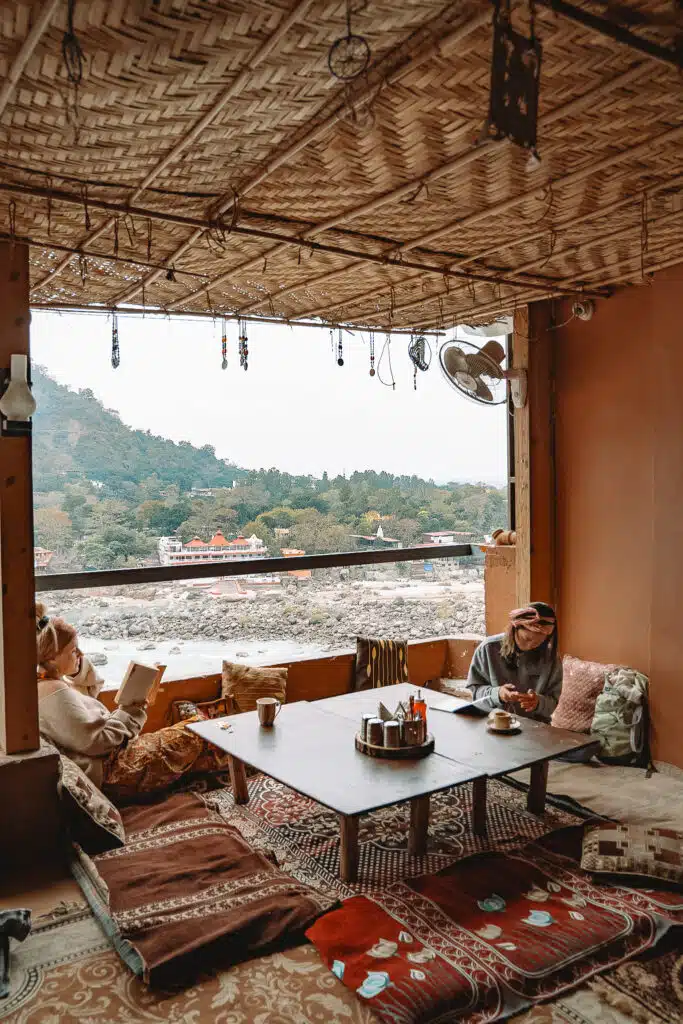 Backpacking India Two people relax in a cozy, open-air cafe with floor cushions and a low table, overlooking a river and mountains—showcasing the warm, inviting atmosphere that often makes travelers ask: Is India Safe For Solo Female Travellers?.