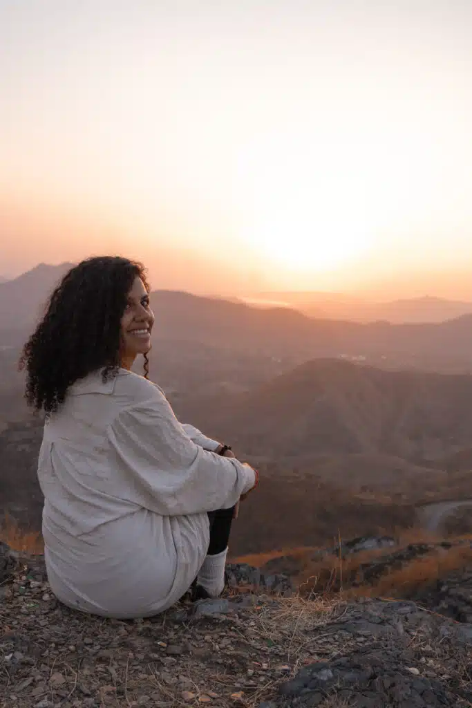 Backpacking India A woman with curly hair sits on a rocky hill, smiling at the camera. She faces a scenic mountain landscape with a warm, glowing sunset in the background—perfect for those wondering, "Is India Safe For Solo Female Travellers?" The scene radiates peace.