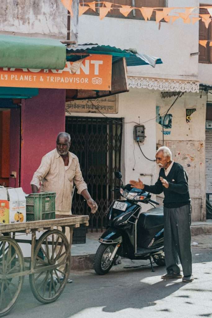 Two elderly men interacting on a street. One man, next to a wooden cart with various items, is wearing a light shirt. The other man, extending his arms, is in a dark sweater beside a parked scooter. They appear to be having a lively conversation about their 2-day Pushkar itinerary.