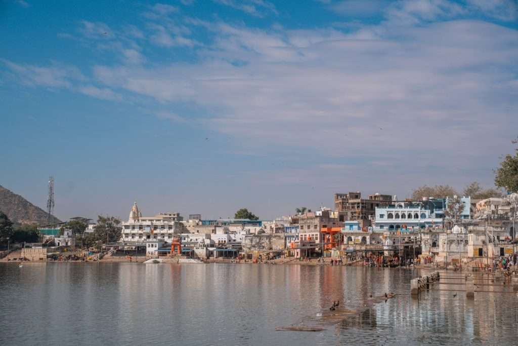 A picturesque waterfront scene of a town with colorful buildings along the shore of a tranquil body of water evokes the question "is Pushkar safe for solo female travelers?" There are various architectural styles, with some structures painted in vibrant blue, white, and orange. The clear sky above adds to the serene ambiance.