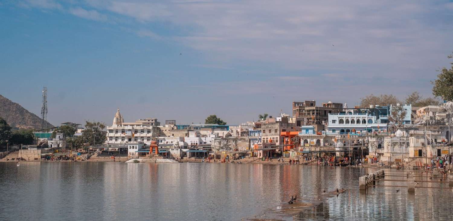 Enjoy a scenic view of a lakeside town on your 1 week Rajasthan itinerary Itinerary, where colorful buildings and temples reflect in the calm water. People gather near the steps leading into the lake under a partly cloudy sky, while hills provide a stunning backdrop.