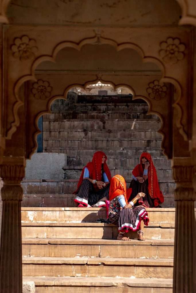 Three women in traditional attire sit and relax on stone steps framed by an ornate archway, reminiscent of a serene moment from a 2-Day Pushkar Itinerary. Two sit upright, while the third lounges with her legs resting on one of the other's lap, creating a casual and serene scene.