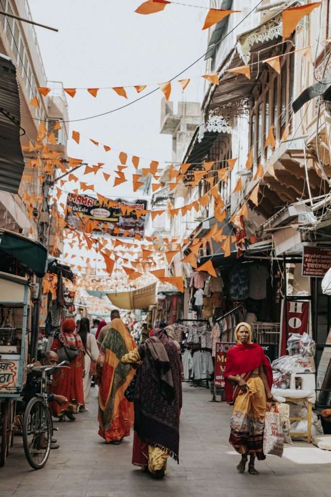 A bustling street market in Pushkar, India with colorful orange flags strung overhead. Several women in traditional sarees walk along the street, past shops filled with various goods, reminiscent of a 2-Day Pushkar Itinerary. The scene is lively with shoppers and vendors engaging in daily activities.