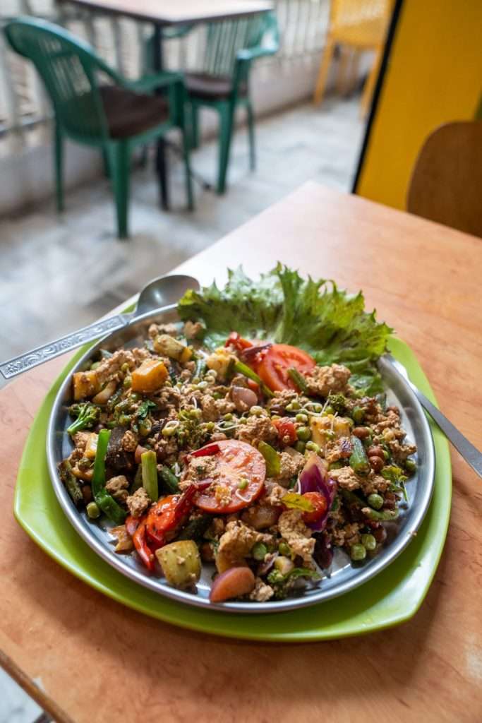 A colorful plate of mixed vegetable salad placed on a wooden table in a casual dining setting. The salad is garnished with lettuce, tomato, potatoes, bell peppers, red onions, beans, and nuts. A spoon and fork are placed beside the plate. In the background, a 2-Day Pushkar Itinerary can be spotted amidst green and yellow chairs around another table. Honey and Spice Cafe Pushkar