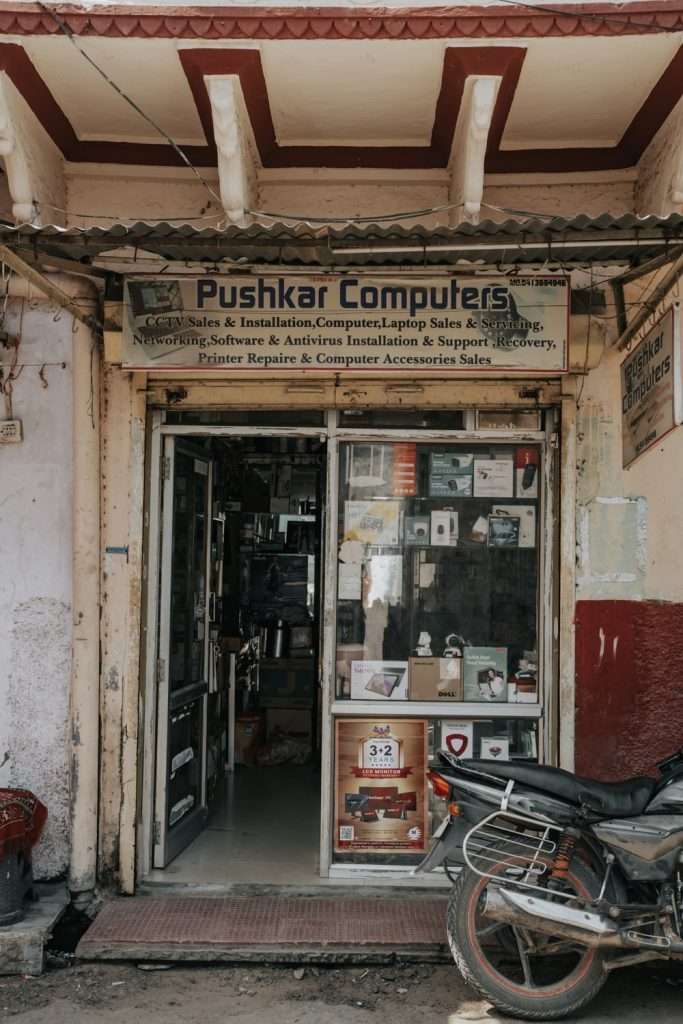 A small computer shop named "Pushkar Computers" with a weathered exterior stands as a hidden gem in this quiet town. The store's entrance is filled with shelves displaying various computer parts and accessories. For anyone on a 2-day Pushkar itinerary, this is an unexpected but intriguing stop, with even a motorcycle parked just outside the shop.