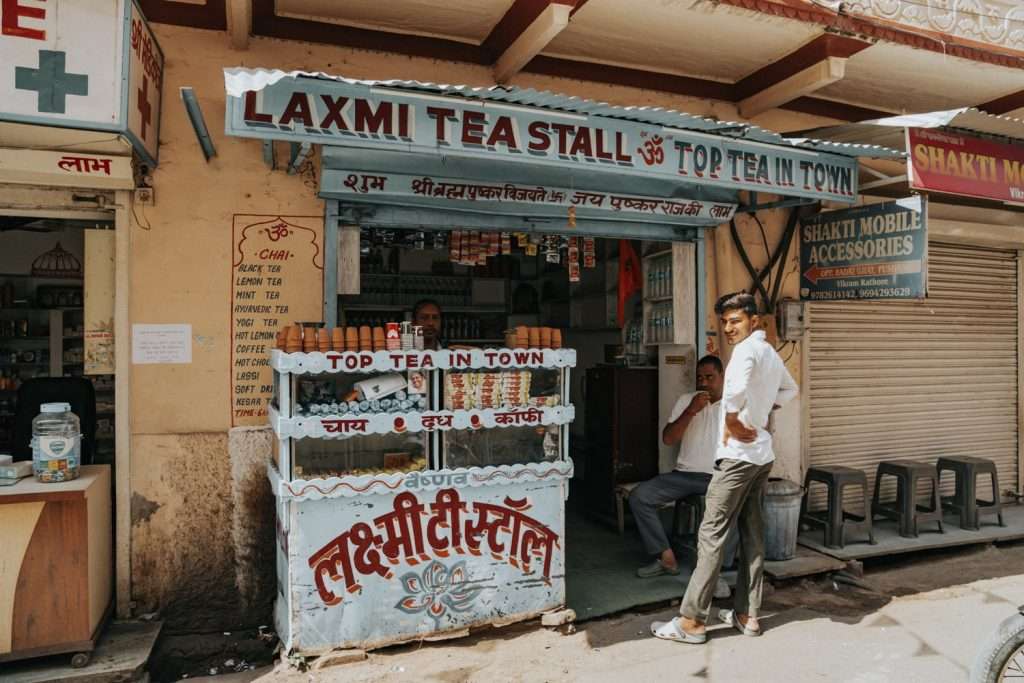 A small street-side tea stall named "Laxmi Tea Stall" with a sign proclaiming "Top Tea in Town." Two men are inside preparing tea while another man stands leaning against the front counter. The stall is adorned with various signs, including one for a 2-Day Pushkar Itinerary, and a neighboring shop is partially visible on the right.