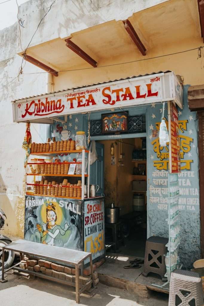 Image of a small tea stall named "Krishna Tea Stall" in India. The shopfront, part of a 2-Day Pushkar Itinerary, is painted with colorful signs advertising tea, coffee, and lemon tea. Terracotta cups are displayed on a shelf. The building appears weathered and is located on a narrow street.