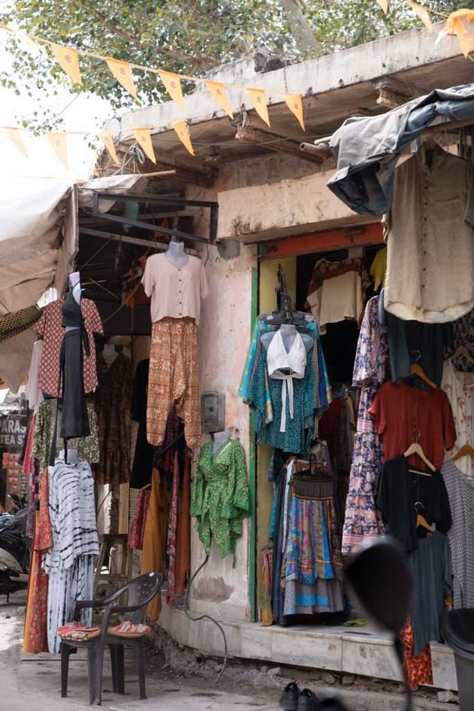 An outdoor clothing shop displays various colorful garments, including dresses, shirts, and skirts. The store is set up on a weathered concrete structure with hanging flags above. Two plastic chairs sit outside, and trees and additional shops can be seen in the background—perfect for a 2-Day Pushkar Itinerary visit.