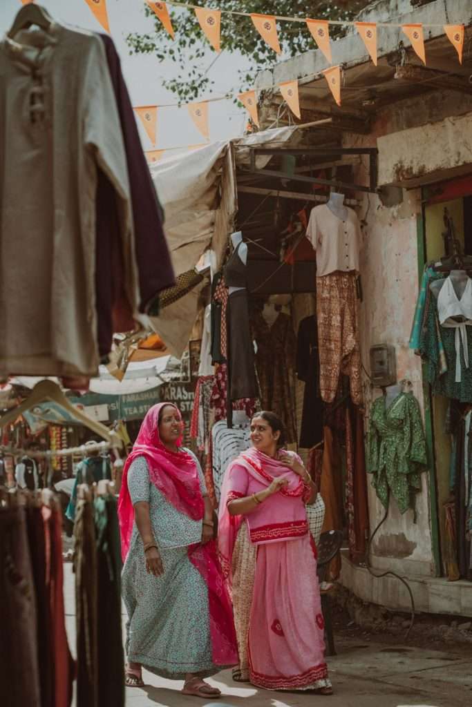 Two women in traditional attire walk through a lively outdoor market, perhaps as part of their 2-day Pushkar itinerary. They wear vibrant pink and red saris while colorful clothing items fill the market stalls around them. Strings of triangular flags are suspended above, with trees visible in the background.