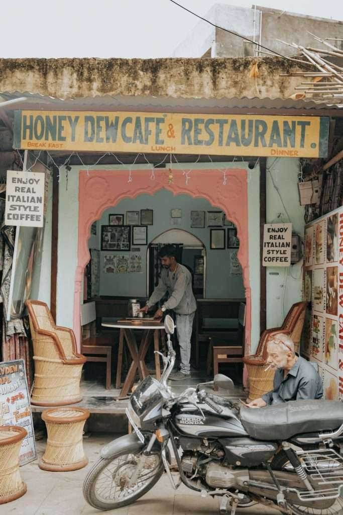 A quaint café named "Honey Dew Café & Restaurant" has a vibrant exterior with a sign displaying its name. A man is setting up wooden tables inside. An elderly man sits outside on a parked motorcycle. Signage promotes "REAL ITALIAN STYLE COFFEE." It's perfect for unwinding after your 2-Day Pushkar Itinerary.