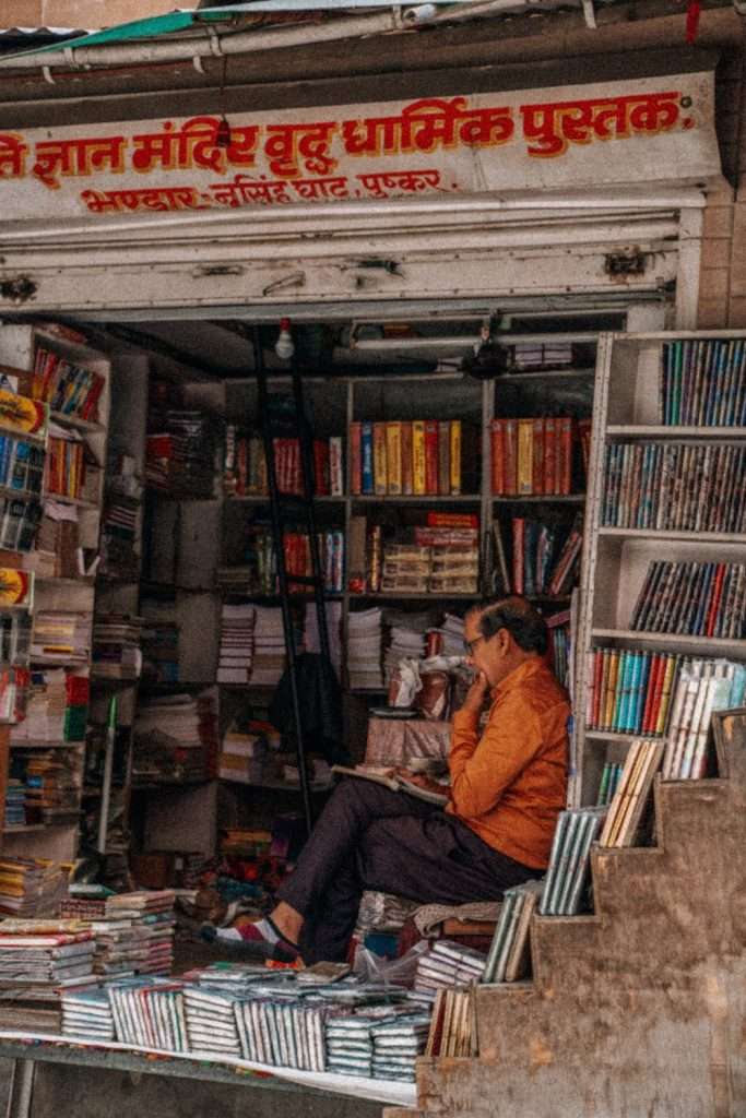 A person in an orange shirt sits inside a small, cluttered bookstore filled with shelves of colorful books. The shop has a sign with Hindi text above the entrance. Perhaps they're planning their 2-day Pushkar itinerary while reading a book or checking their phone.