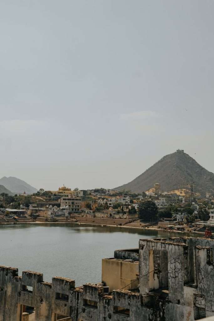 A scenic view of a town by a lake from Lauras Cafe with a mountainous backdrop. Buildings line the shore, and a larger hill with structures on top is visible in the background. The sky is clear and light blue. Weathered structures in the foreground hint at a historical setting, reminiscent of exploring an adventurous 2-Day Pushkar Itinerary.