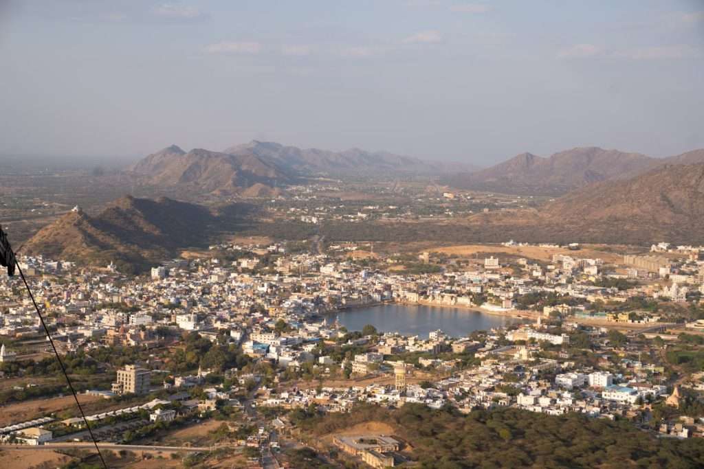 Aerial view of a sprawling city surrounded by hills and mountains. In the center of the city, there is a large lake, with numerous buildings and green spaces around it, perfect for those planning a 2-day Pushkar itinerary. The sky is clear with soft sunlight casting shadows on the landscape.