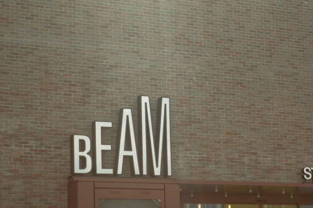 The image shows a red brick wall with a sign that reads "BEAM" in large, white, uppercase letters. The sign is situated on top of a storefront—in what could be one of the popular restaurants in Hertford—displaying a modern and minimalistic architectural style. - Beam Theatre Hertford