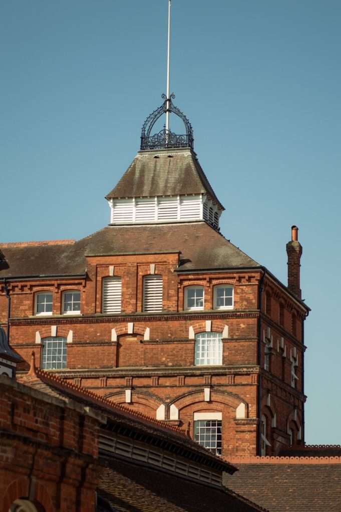 A tall, historical red-brick building with several stories, featuring rows of windows, decorative brickwork, and a distinctive rooftop structure with a flagpole. Set against the clear blue sky that enhances its architectural details, it stands as one of the notable landmarks among in Hertford.