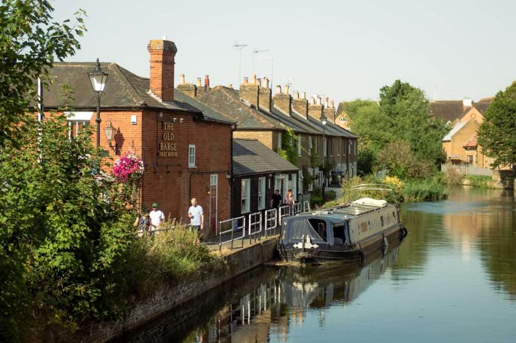 A serene canal scene with a long, narrow boat moored alongside a row of charming brick houses. A pub named "The Old Barge" is visible, with people walking by the waterside and lush greenery surrounding the area—one of the delightful spots among many of the best things to do in Hertford.