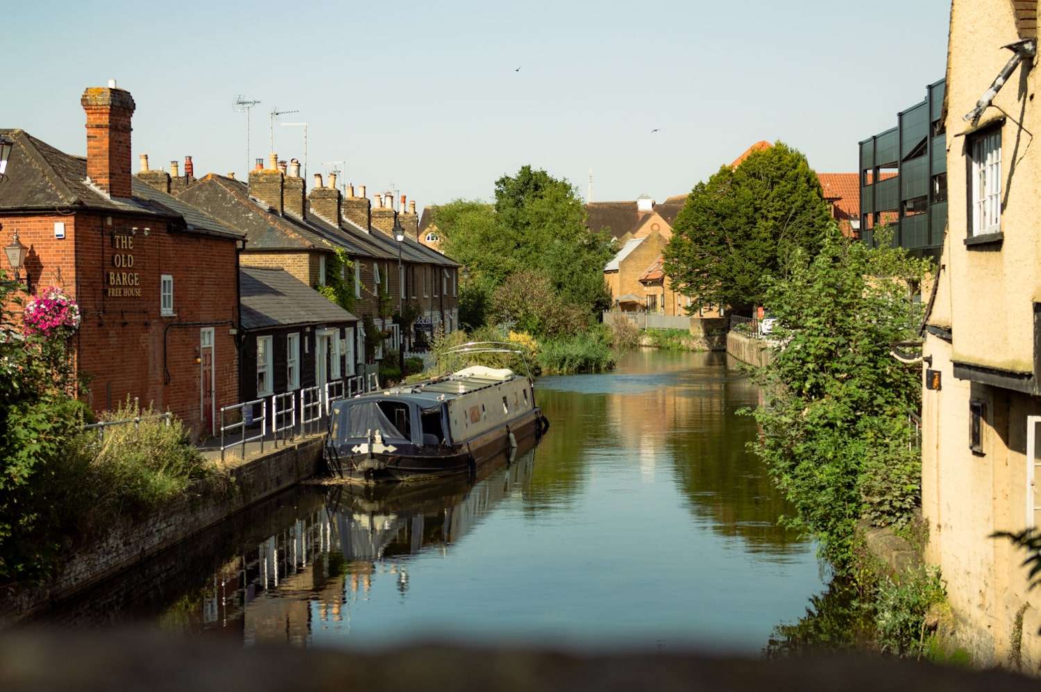 A narrow canal with a white houseboat is flanked by old brick houses and lush greenery on both sides. Several chimneys and rooftops are visible under a clear blue sky. In the background, more buildings and trees are seen, creating a serene and picturesque scene—one of many things to do in Hertford.