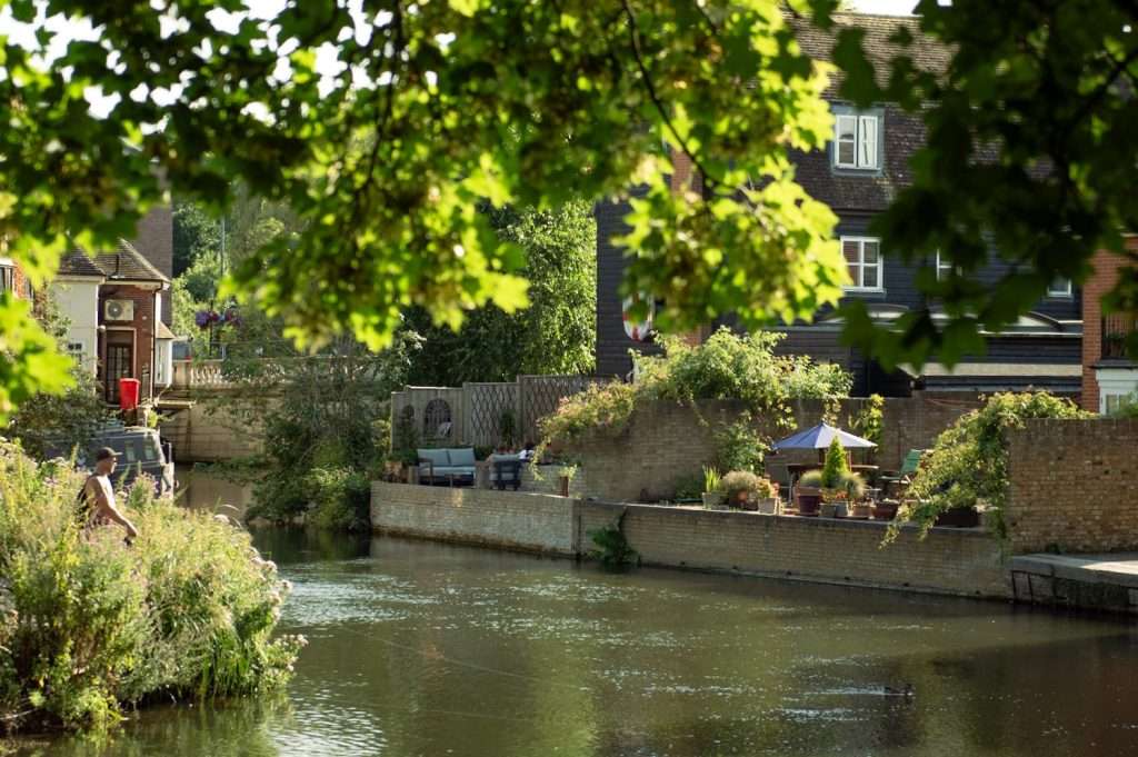 A serene riverside scene with leafy tree branches framing the top. A person is fishing on the riverbank surrounded by lush greenery. Across the water, there's a patio with outdoor furniture and potted plants next to charming brick houses—one of the delightful things to do in Hertford.