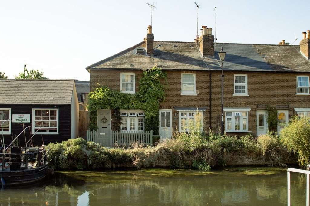 A serene riverside scene in Hertford with a row of charming, rustic brick houses partially covered in ivy. The houses have chimneys and TV antennas on their roofs. A few boats are moored by the edge of the calm river, enhancing the tranquil atmosphere.