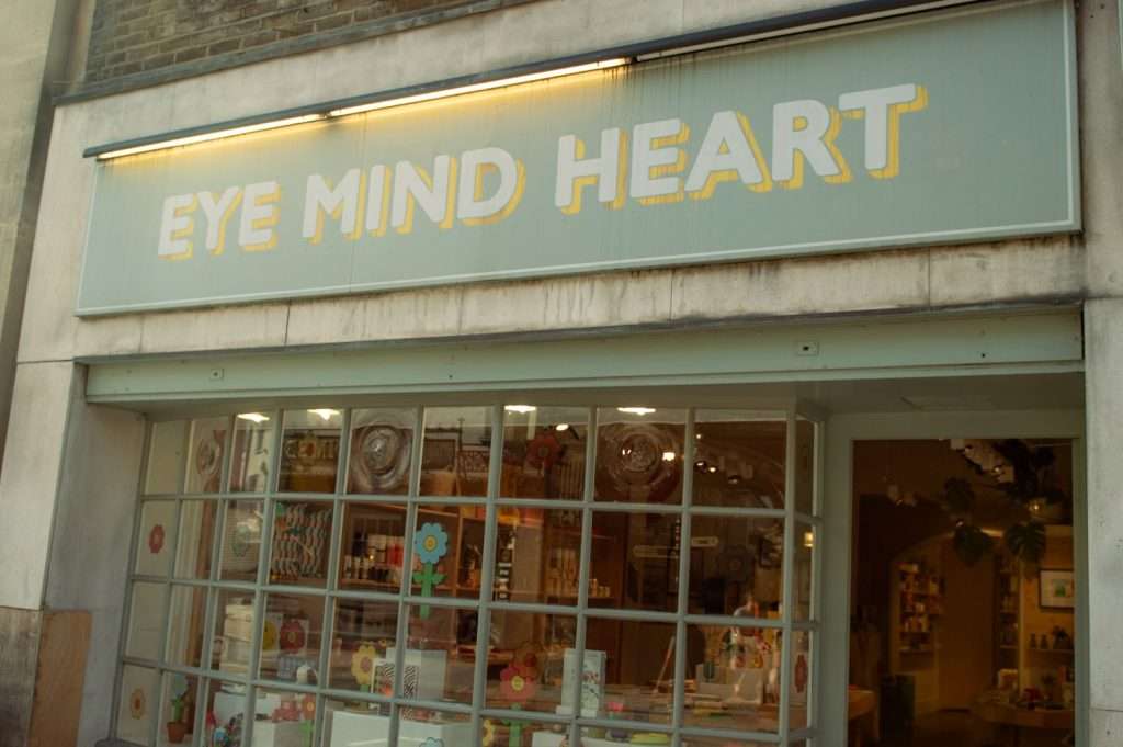 A storefront with large windows displaying various items. A light blue sign above the entrance reads "EYE MIND HEART" in white, bold letters. Inside the store, the shelves are filled with colorful products and decorations. Among the things to do in Hertford, this charming shop stands out despite its slightly weathered exterior.