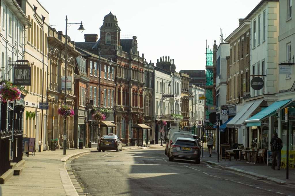 A narrow, slightly curved street lined with historical buildings on a sunny day offers a glimpse of the many things to do in Hertford. The mix of shops, cafes, and eateries adorned with hanging flower baskets is inviting. Few cars are parked along the street, and a couple of people are walking.