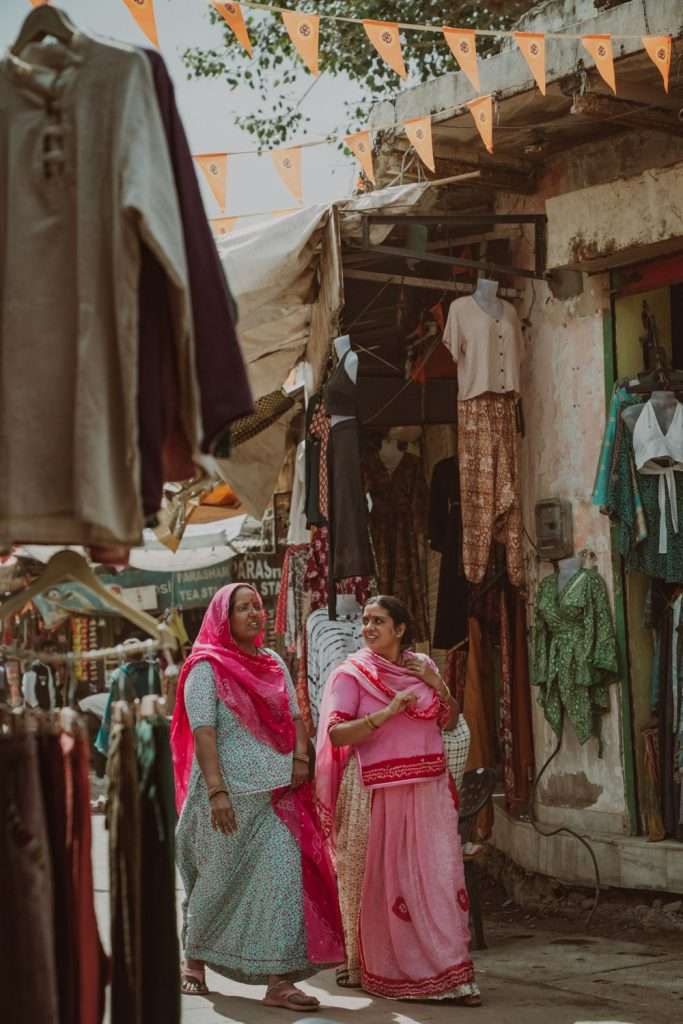 Two women in colorful traditional Indian clothing stroll through a bustling street market, vibrant stalls and hanging garments all around, with orange flags strung overhead—an authentic scene from a perfect 2-Day Pushkar Itinerary.