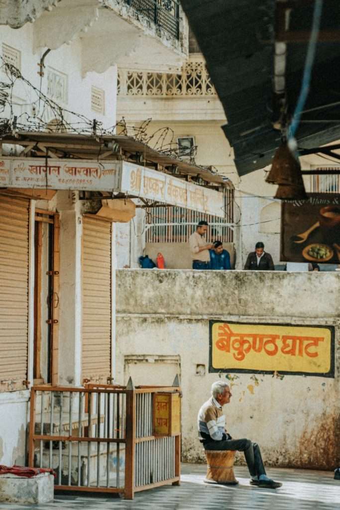An elderly man sits alone on a bench in front of closed shops with faded Hindi signs, in a quiet street that hints at the slower pace outside a bustling 2-Day Pushkar Itinerary, all bathed in warm Indian sunlight.