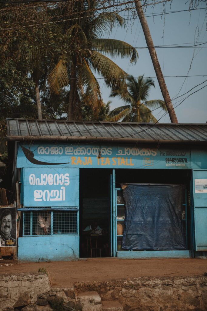 A blue tea stall named "Raja Tea Stall" with signage in Malayalam and English, perfect for a 1-week Kerala itinerary, sits beside a road lined with palm trees. A portrait and phone numbers are displayed on the wall.