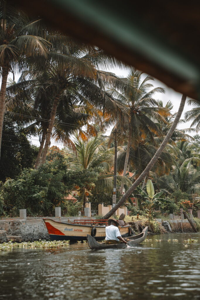 A person paddles a small canoe on a calm river, surrounded by lush green palm trees—a perfect scene from a 1-week Kerala itinerary. A colorful boat is docked on the riverbank in the background under the tropical foliage.