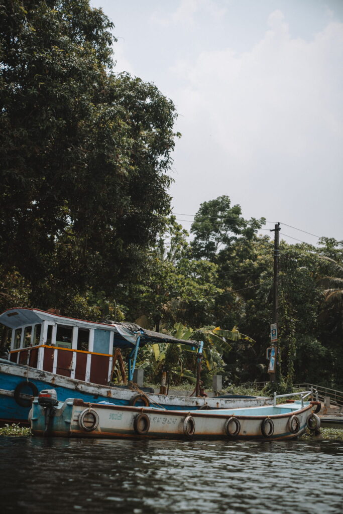 A small, weathered blue and white boat floats on calm water near a lush, green shoreline with tall trees and utility poles under a cloudy sky—an idyllic scene for any 1-week Kerala itinerary.