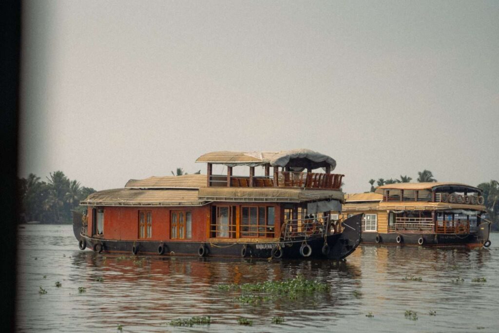 Two traditional houseboats with wooden exteriors float on calm water, surrounded by floating greenery—an idyllic scene perfect for a 1-week Kerala itinerary. Palm trees and hazy skies in the background enhance the tropical, tranquil setting.