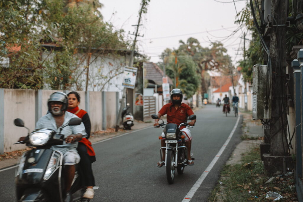 People ride motorcycles and scooters on a narrow street lined with houses and trees—a vibrant scene you might encounter during your 1-week Kerala itinerary, with riders and pedestrians adding to the local charm.