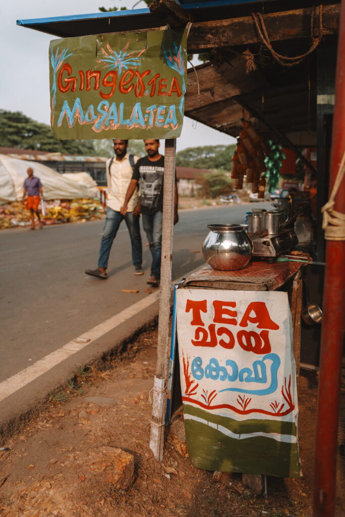 A small roadside tea stall with colorful hand-painted signs advertising ginger tea and masala tea—an ideal stop during your 1-week Kerala itinerary. Two men walk by as metal teapots and cups glint on the counter.