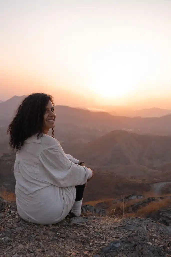 A woman with curly hair sits on a rocky hilltop at sunset, looking over her shoulder and smiling. The scene, reminiscent of the tranquil landscapes near Udaipur, might leave you wondering: Is Udaipur worth visiting for views like this?.