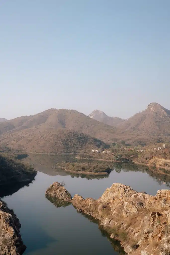 A calm river winds through rocky terrain, reflecting brown hills and a clear blue sky. Small houses rest at the base of the hills, evoking a serene, untouched landscape—just one scene that answers the question: Is Udaipur worth visiting?.