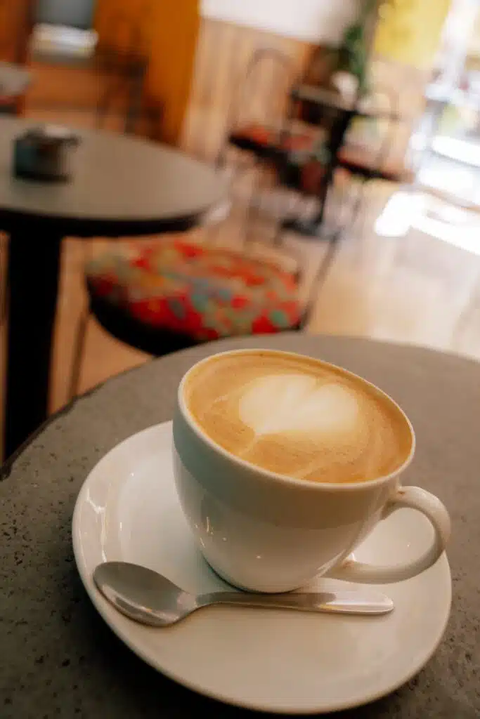 A cup of cappuccino with heart-shaped latte art sits on a white saucer atop a café table. In the background, empty tables glow in warm, natural light—perfect for reflecting on questions like "Is Udaipur Worth Visiting?.