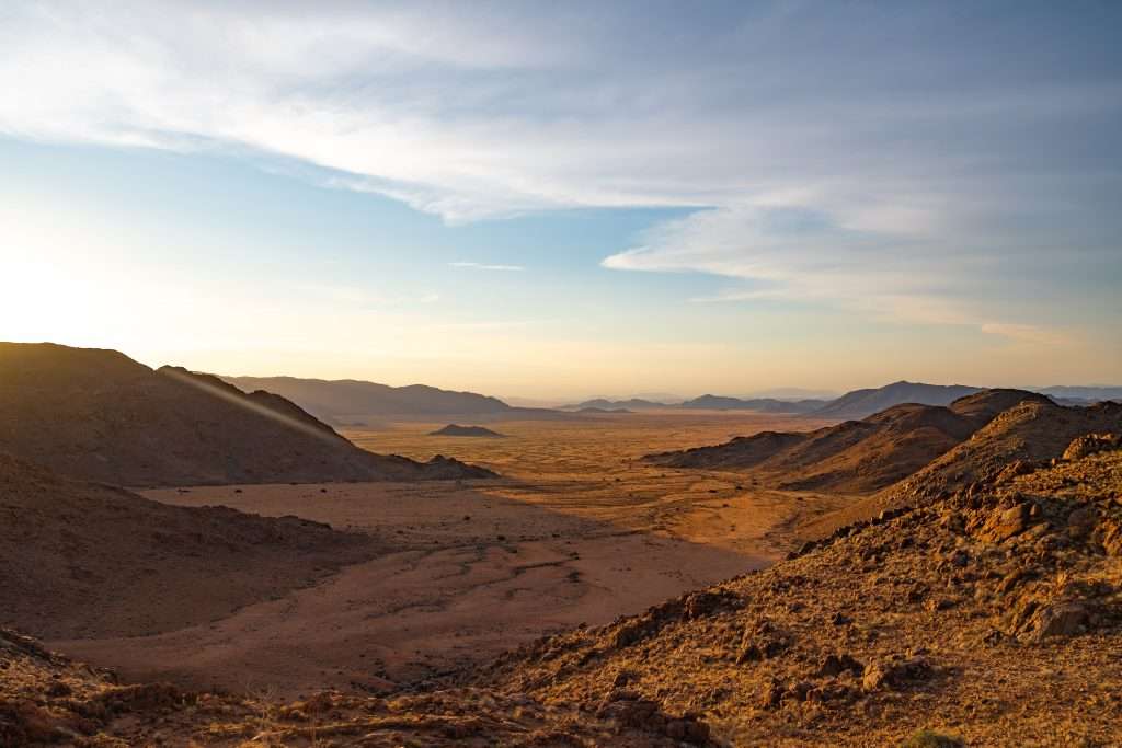 Is Namibia worth visiting? Absolutely. Picture a vast desert landscape with rocky mountains silhouetted in the foreground and middle distance. The terrain blends rugged rocks with sandy plains under a clear sky adorned with scattered clouds, as the setting sun casts a warm glow over this stunning scenery. - The Namib Desert 