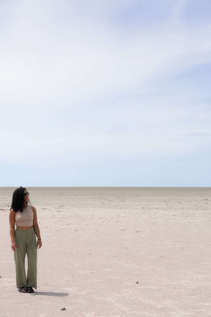 A woman with curly hair and sunglasses stands on a vast, empty beach under a bright blue sky. She is wearing a white sleeveless top and green pants. The sand stretches far into the distance, meeting the horizon with a light, cloud-streaked sky above- Etosha National Park Salt Pan Is Namibia worth visiting? Absolutely!