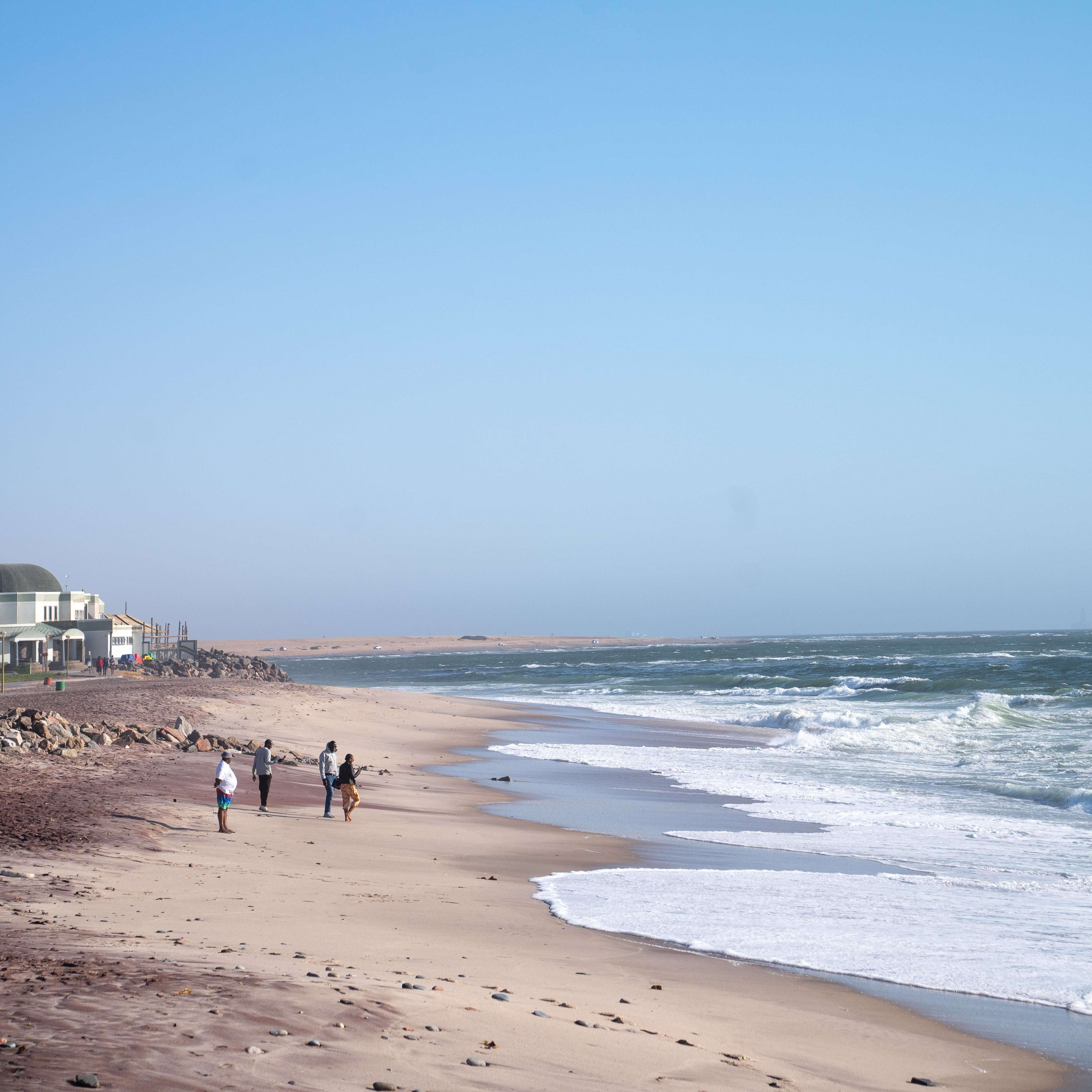 Two people and a dog walk along a vast, sandy beach with gentle waves, evoking the question: "Is Namibia worth visiting?" A few houses are visible on the left side near the shore. The sky is clear and blue, stretching above the tranquil sea.