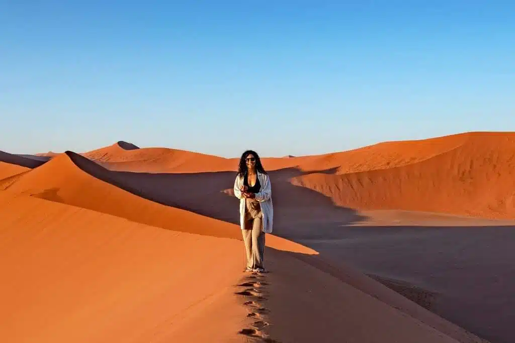 A person stands atop a tall sand dune in Namibia's vast desert landscape, holding a camera. The dunes feature varying shades of orange under a clear, blue sky. The person wears a light-colored outfit and sunglasses, appearing engaged in the serene scenery. Is Namibia safe for solo female travelers?