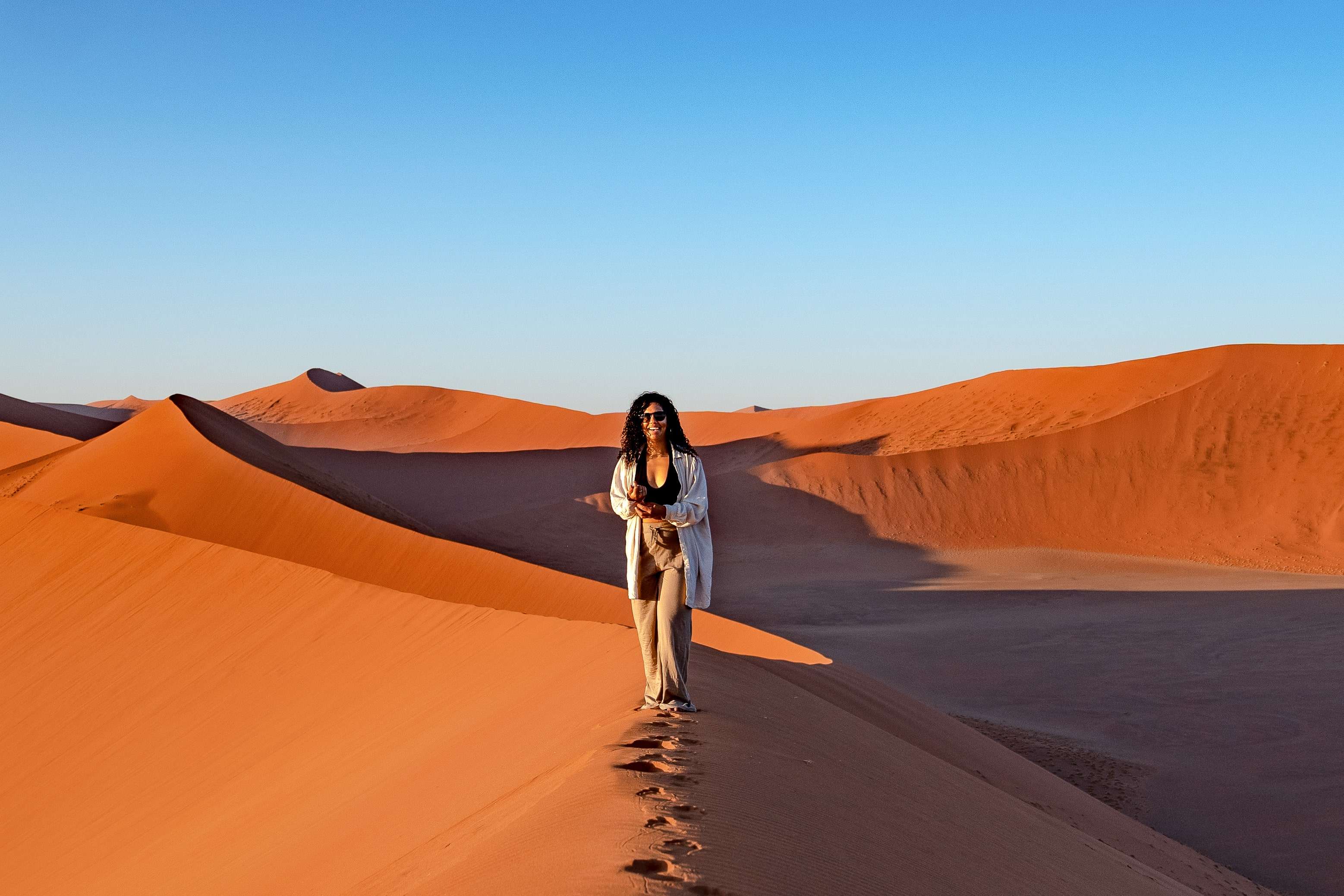 A person stands atop a tall sand dune in Namibia's vast desert landscape, holding a camera. The dunes feature varying shades of orange under a clear, blue sky. The person wears a light-colored outfit and sunglasses, appearing engaged in the serene scenery. Is Namibia safe for solo female travelers?