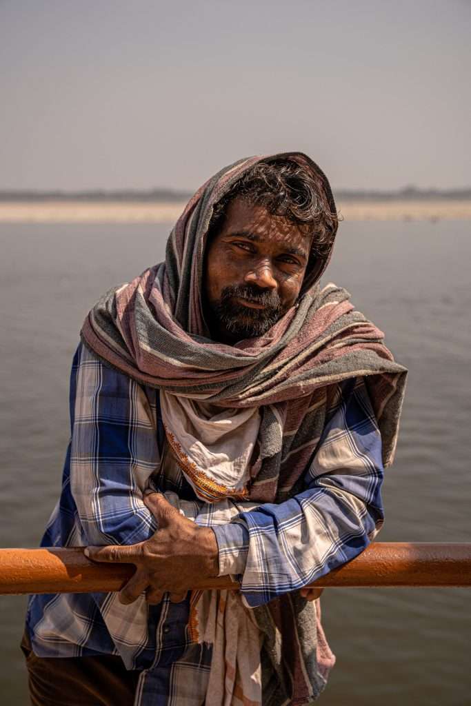 A man is leaning against a wooden railing, with a calm river and sandy shore in the background. He is wearing a blue plaid shirt and has a shawl draped over his shoulders and head. The man is looking directly at the camera, with a slight smile on his face, as if pondering, "Is Varanasi worth visiting?