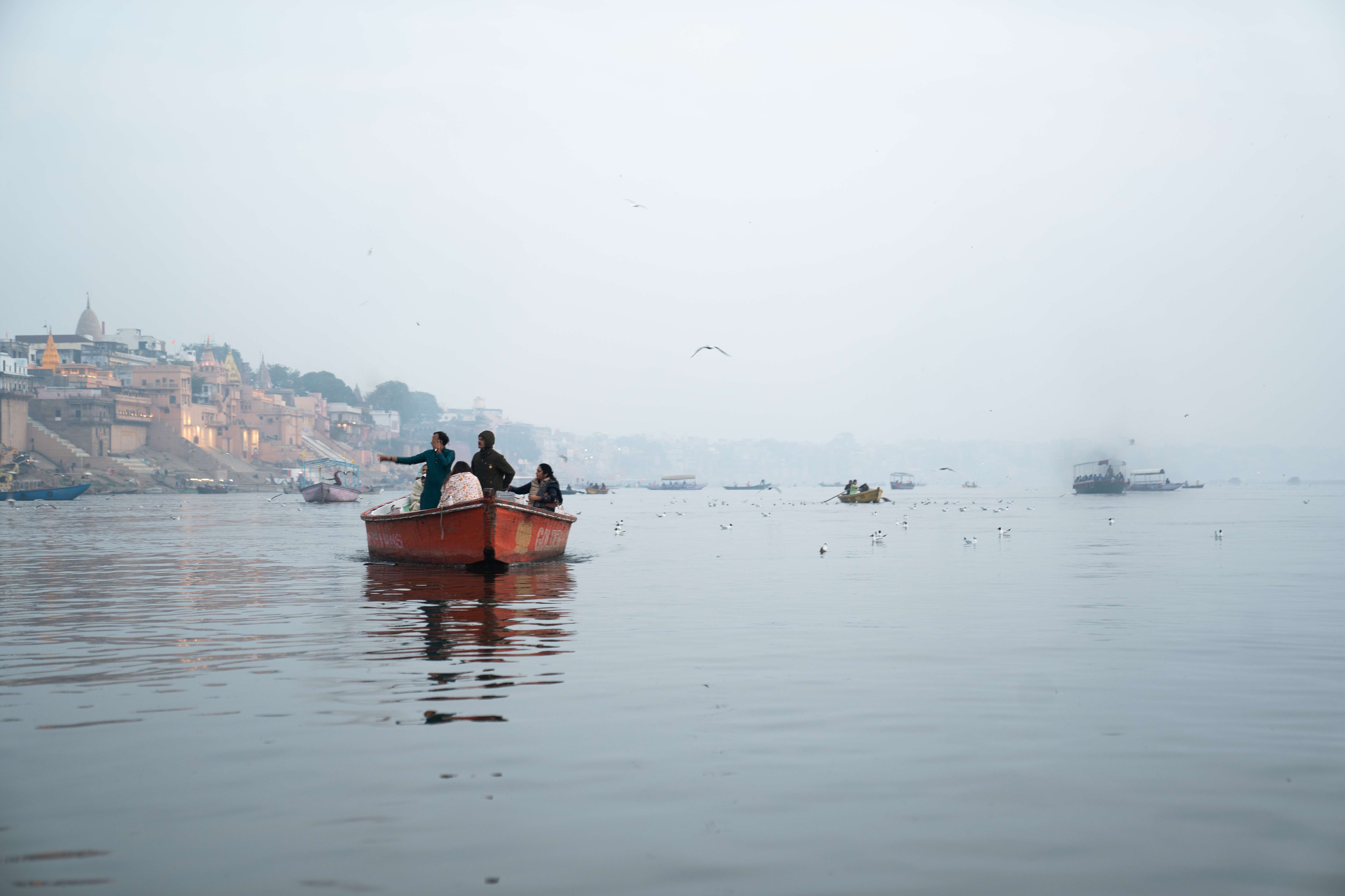 A red boat with several people on board floats on a calm river beside an ancient city with historic buildings and temples, reminiscent of Varanasi. Birds fly above the water, and additional boats are scattered in the distance, all under a hazy sky. Is Varanasi worth visiting for scenes like this?