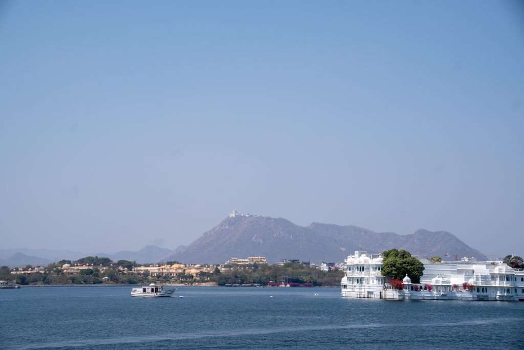 A serene lake with calm blue water is seen in the foreground. A small boat sails on the lake. The background features a cityscape and distant mountains under a clear blue sky. On the right, there's a large white building by the water.