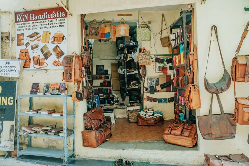 A small shop in Udaipur showcases various handcrafted leather goods, including bags, purses, and book covers. Leather items hang on the walls and are displayed on shelves. A signboard reads "KGN Handicrafts". Outside, a step ladder holds additional items. Is Udaipur worth visiting? Absolutely!